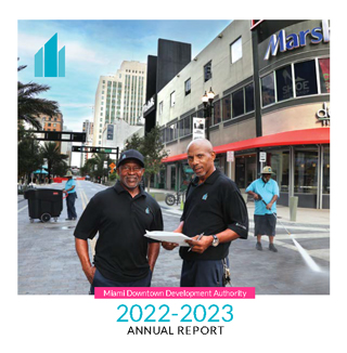 Cover image of the 2022-2023 Miami DDA Annual Report. Two brown men wearing black shirts. THe man on the left wearing a black baseball cap. The other man bald. Both men are members of the Enhanced Services Team are standing in the center of Flagler St. taking a picture before continuing to work.